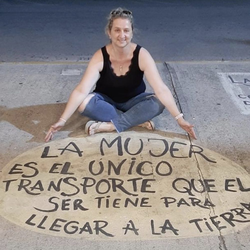 Claudia Amuchastegui sentada junto a una intervención callejera que dice: la mujer es el único transporte que el ser tiene para llegar a la tierra
